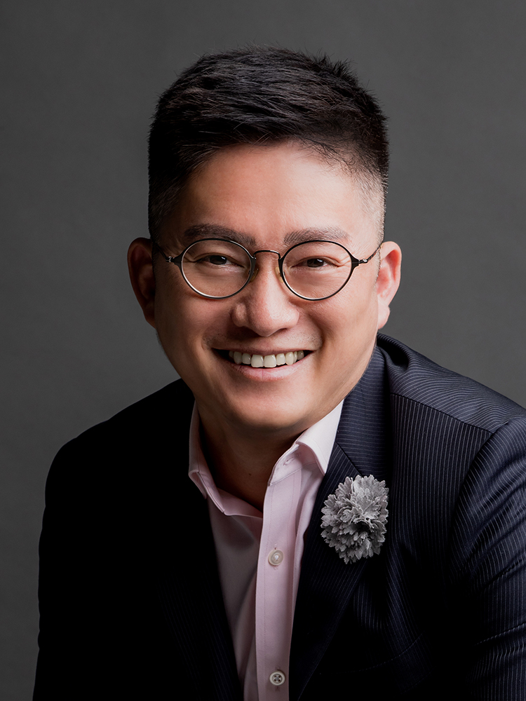 一位戴着眼镜、身穿西装外套和粉色纽扣衬衫的黑发男子正在拍照